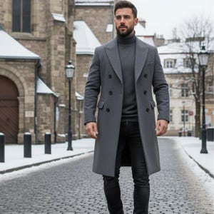 Könnte beinhalten: Ein grauer, doppelreihiger Mantel mit fallendem Revers und einem passenden grauen Rollkragenpullover. Die Person trägt eine schwarze Jeans und geht auf einer Kopfsteinpflasterstraße. Der Hintergrund umfasst Gebäude und Straßenlaternen.