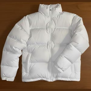 Könnte beinhalten: Eine weiße Steppjacke mit hohem Kragen und durchgehendem Reißverschluss. Die Jacke hat ein gestepptes Design und zwei Seitentaschen. Die Ärmel sind lang und die Jacke scheint aus einem glatten, wasserabweisenden Material zu sein. Die Jacke liegt flach auf einer Holzoberfläche.