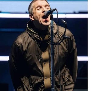 Può includere: Un uomo canta in un microfono sul palco. Indossa una giacca verde scuro con una striscia color beige sul davanti. La giacca ha un cappuccio ed è abbottonata. Lo sfondo è scuro con luci blu e bianche.