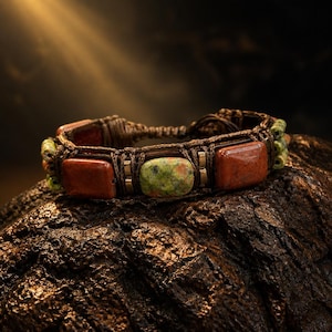May include: A bracelet featuring rectangular reddish-brown stones and a central green and speckled stone, all woven into a dark brown cord. The design includes small green accent beads and metallic spacers, creating a structured, multi-textured band.