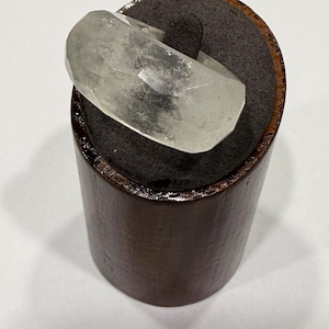 Può includere: Un anello trasparente sfaccettato realizzato in un materiale cristallino traslucido. L'anello presenta molteplici superfici geometriche piatte che catturano la luce, creando un effetto scintillante. È esposto su un supporto cilindrico scuro.