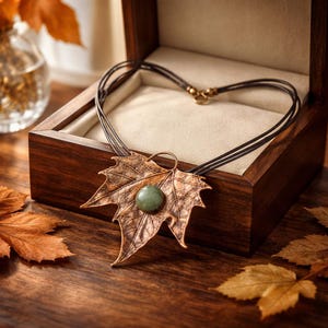 Pode incluir: Um colar com pingente de folha de cor bronze com uma pedra verde, apresentado numa caixa de joias de madeira. O colar tem um cordão preto de vários fios. A caixa está aberta, revelando um interior creme.