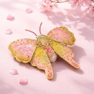 Può includere: Spilla a farfalla artigianale con ali ricamate rosa e gialle, adornata con perline e una gemma centrale. La spilla è posta su uno sfondo rosa tenue con petali e fiori di ciliegio sparsi.