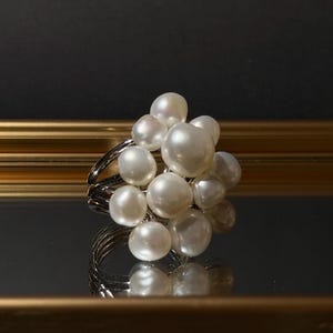 Op de afbeelding: Een ring met een cluster van meerdere witte, glanzende parels in onregelmatige vormen, gerangschikt in een ronde formatie. De parels zijn bevestigd op een zilverkleurige metalen band die bestaat uit meerdere dunne, parallelle draden.
