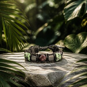May include: A woven cord bracelet featuring a central dark red polished stone, flanked by two smooth silver-toned square beads and two textured green and black speckled stone segments, finished with an adjustable sliding knot closure mechanism.