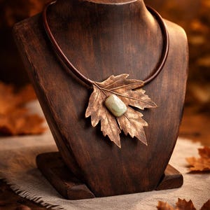 Pode incluir: Colar com pingente de folha em tom de cobre com uma pedra verde clara no centro, exibido em um busto de madeira. O colar tem um cordão de couro marrom. O busto está sobre uma base de madeira, com folhas de outono ao fundo.