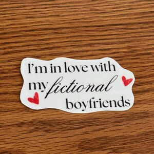 May include: A white sticker with the text "I'm in love with my fictional boyfriends" in black script. Two small red hearts are on the sticker. The sticker is on a wooden surface.