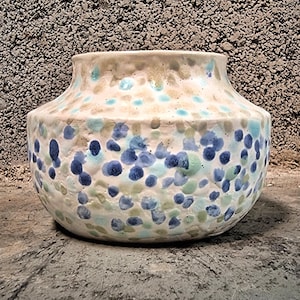 Puede incluir: Un jarrón de cerámica con un cuerpo ancho y redondeado y un borde ligeramente ensanchado. El jarrón está decorado con un patrón de puntos azules, verdes y blancos. El fondo es una superficie gris texturizada.