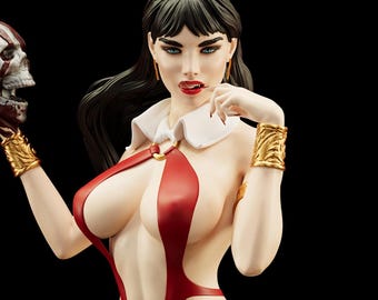 May include: A detailed figurine of a woman with long black hair, blue eyes, and red lipstick. She wears a red and white outfit with gold arm cuffs and earrings. She holds a skull in her left hand. The background is black.