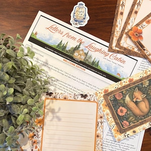 May include: A printed publication titled Letters from the Laughing Cabin Welcome Edition, featuring a cabin and forest illustration, accompanied by floral-bordered stationery, a sleeping fox card, and a lantern sticker with the text Be A Light For All To See.