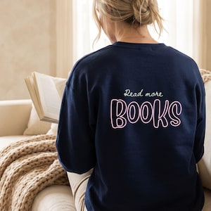 Könnte beinhalten: Marineblaues Sweatshirt mit dem Text "Read more BOOKS" in Weiß und Pink. Die Person, die das Sweatshirt trägt, liest ein Buch. Der Hintergrund zeigt ein beiges Sofa und ein Fenster.