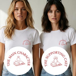 Könnte beinhalten: Zwei weiße kurzärmelige T-Shirts mit roten Grafikdrucken auf der Brust. Ein Shirt zeigt ein offenes Buch mit einem Diamantring und dem Text THE MAIN CHARACTER. Das andere Shirt zeigt ein offenes Buch mit zwei Weingläsern und THE SUPPORTING CAST.