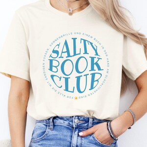 Könnte beinhalten: Ein cremefarbenes Kurzarm-T-Shirt mit einer kreisförmigen blauen Grafik und dem Text SALTY BOOK CLUB in der Mitte. Der umgebende Text lautet FÜR ALLE DIE MIT SANDIGEN FÜSSEN, SONNENBRILLE UND EINEM BUCH IN DER HAND AM GLÜCKLICHSTEN SIND.