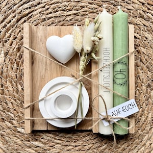 Könnte beinhalten: Ein Holztablett enthält ein weißes Herz, einen kleinen Strauß Trockenblumen und einen weißen Kerzenhalter. Zwei Kerzen sind enthalten, eine cremefarbene mit Text und eine grüne mit der Zahl 40. Die Kerzen sind mit Bindfaden zusammengebunden, und ein Etikett lautet "AUF EUCH."