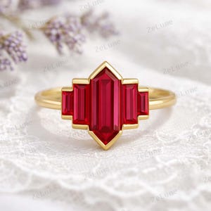 Puede incluir: Un anillo de oro con un diseño geométrico, con una gema roja rectangular central y gemas rojas rectangulares más pequeñas. El anillo tiene un acabado pulido. El fondo es blanco.