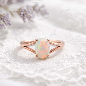 Peut inclure: Bague en or rose ornée d'une opale ovale. L'opale présente un jeu de couleurs, sertie dans une monture à fente. La bague est présentée sur une surface en dentelle blanche. Le texte "ZeLume" est visible.