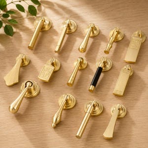 May include: A collection of twelve brass furniture pulls and drop handles in various shapes, including cylindrical, rectangular, and tapered designs. Some feature engraved geometric patterns, while one handle includes a black cylindrical accent.