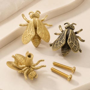 May include: Three bee-shaped cabinet knobs made of brass with a metallic finish. Two knobs are shown from the top, displaying detailed wing patterns and segmented bodies. One knob is shown from the side, revealing the threaded mounting post.