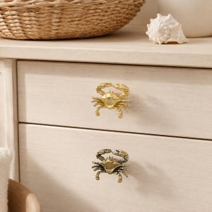 Puede incluir: Dos pomos de cajón metálicos con forma de cangrejo fijados a un mueble de madera clara. El pomo superior tiene un acabado dorado brillante, mientras que el inferior presenta un acabado en latón antiguo más oscuro con patas y pinzas detalladas.