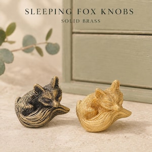 May include: Two solid brass knobs shaped like sleeping foxes. One knob has a dark, antiqued finish, while the other has a bright, polished gold-tone finish. The text SLEEPING FOX KNOBS SOLID BRASS is visible at the top of the image.