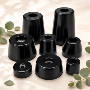 May include: A collection of eight black, tapered, cylindrical rubber feet or bumpers of varying heights and widths. Each piece features a smooth, glossy finish and a central circular opening with a metallic insert visible at the base of the hole.
