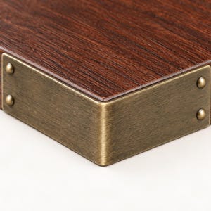 May include: A dark brown wood corner piece featuring a brushed brass metal corner guard. The metal guard is L-shaped and secured to the wood with four small, rounded brass rivets, two on each side of the corner, providing a reinforced edge.