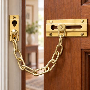 May include: A polished brass door security chain lock installed on a wooden door and frame. The mechanism consists of a rectangular mounting plate with a sliding track, a chain with oval links, and a separate catch plate with a keyhole-shaped slot.