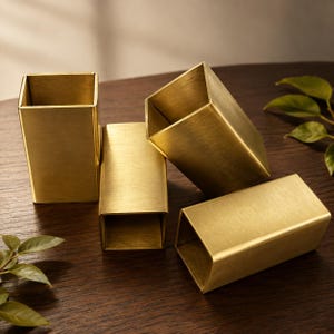 Könnte beinhalten: Vier rechteckige Behälter aus gebürstetem Messing. Jeder Behälter verfügt über einen hohlen Innenraum und eine glatte, goldfarbene Metalloberfläche. Die Objekte sind in einer Gruppe angeordnet, wobei einige aufrecht stehen und andere liegen.