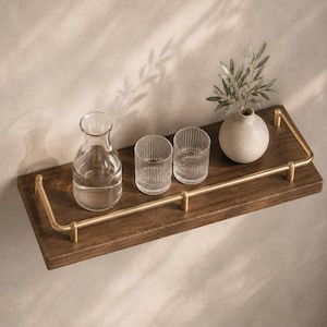May include: A rectangular dark brown wooden wall shelf featuring a brushed gold metal railing along the front and sides. The shelf holds a clear glass carafe, two ribbed glass tumblers, and a small beige ceramic vase with green foliage.