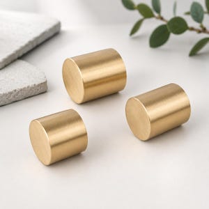 May include: Three cylindrical cabinet knobs with a brushed gold finish. Each knob features a smooth, flat circular face and a solid, uniform metal body. The hardware has a metallic texture and a consistent golden tone throughout the set.
