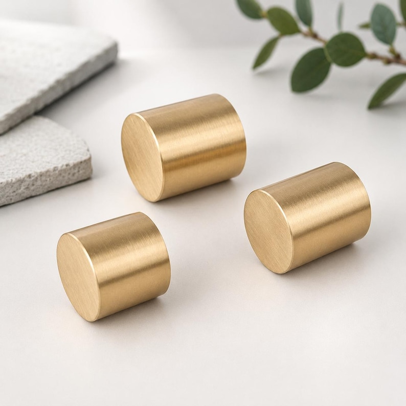 May include: Three cylindrical cabinet knobs with a brushed gold finish. Each knob features a smooth, flat circular face and a solid, uniform metal body. The hardware has a metallic texture and a consistent golden tone throughout the set.