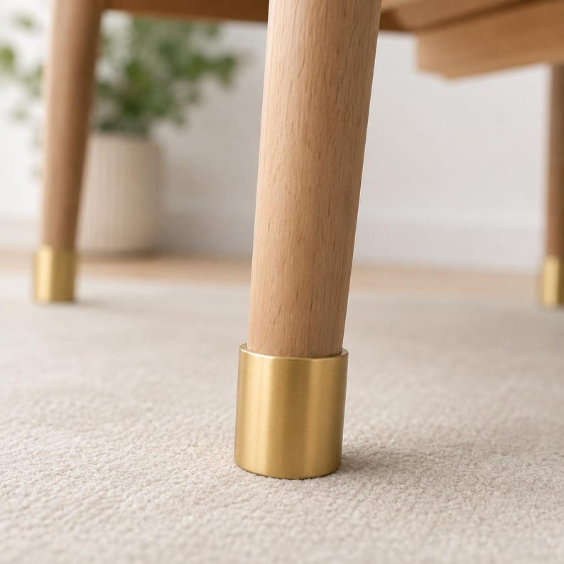 May include: A set of light-colored wooden furniture legs, each featuring a smooth, cylindrical brass-toned metal cap at the base. The legs are tapered and stand upright, showcasing the natural wood grain texture and a polished metallic finish.