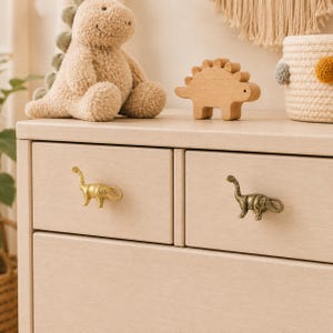 Pode incluir: Dois puxadores de gaveta de metal em forma de dinossauro fixados numa cómoda de madeira clara. Um puxador tem um acabamento dourado brilhante e o outro um acabamento em bronze mate. Ambos os puxadores têm a forma de dinossauros saurópodes.