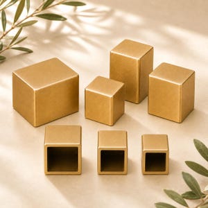 Könnte beinhalten: Eine Sammlung von sechs goldfarbenen Metallblöcken in verschiedenen Größen. Drei der Blöcke sind massive Würfel, während die anderen drei hohle, an den Enden offene rechteckige Prismen sind. Jedes Stück hat eine glatte, matte Oberfläche.