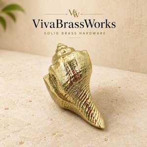 May include: A gold-colored brass cabinet knob shaped like a seashell with a textured surface. The text VivaBrassWorks SOLID BRASS HARDWARE is visible above the object. The knob features a spiral top and a tapered base with metallic finish.