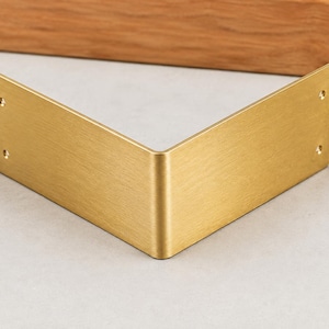 Può includere: Una staffa per mensola in metallo a forma di V con finitura oro spazzolato. Il supporto presenta un design liscio e angolato con due piccoli fori circolari per le viti su ciascuna delle due estremità verticali piatte.