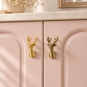 May include: Two gold-colored metal cabinet knobs shaped like deer heads with antlers. The knobs are mounted on two light pink cabinet doors with arched panel detailing. The deer heads feature detailed facial features and textured antlers.
