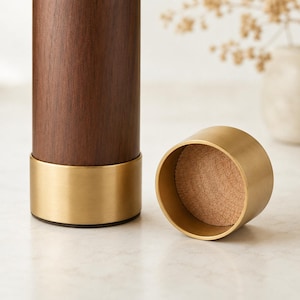 May include: A cylindrical dark brown walnut wood container with a smooth brushed brass metal base. Beside it sits a matching cylindrical brass cap with a circular cork insert visible on the interior, showcasing a clean and functional design.