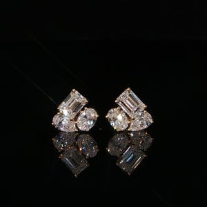 May include: A pair of stud earrings featuring a cluster of three clear, faceted stones in emerald, oval, and pear cuts. The stones are set in a metallic frame and reflect light against a solid black background, creating a symmetrical, brilliant appearance.