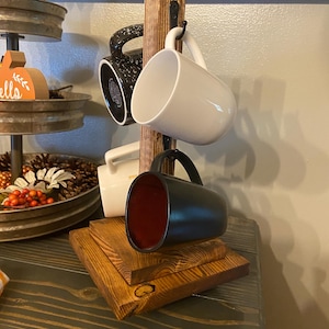 May include: A wooden mug rack with four mugs hanging from it. The mugs are black, white, and speckled. The rack is made of stained wood and sits on a wooden base. The sign above the rack says "UNTIL IT'S TIME FOR WINE."