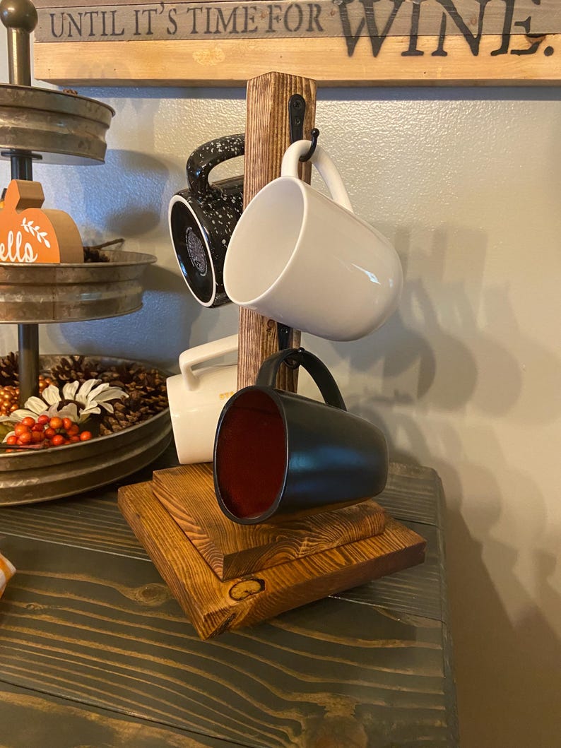 May include: A wooden mug rack with four mugs hanging from it. The mugs are black, white, and speckled. The rack is made of stained wood and sits on a wooden base. The sign above the rack says "UNTIL IT'S TIME FOR WINE."