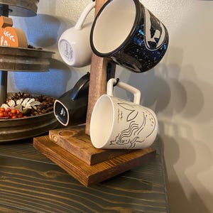 May include: A wooden mug rack with four mugs. The mugs are white, black, and speckled with various designs. The rack is made of stained wood and has a rustic appearance. The mugs are hanging from metal hooks.