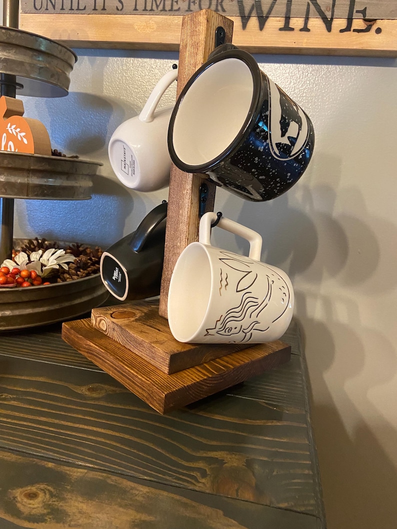 May include: A wooden mug rack with four mugs. The mugs are white, black, and speckled with various designs. The rack is made of stained wood and has a rustic appearance. The mugs are hanging from metal hooks.