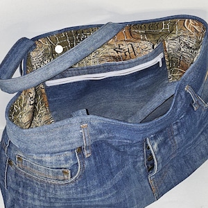 May include: A blue denim tote bag with a camouflage patterned interior. The bag features a zipper closure, a front pocket, and denim handles. The bag is made from recycled denim jeans.