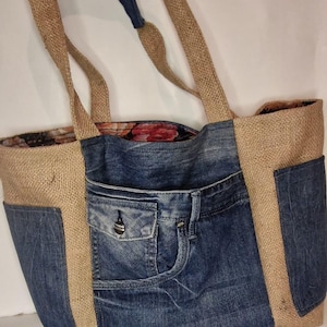Puede incluir: Un bolso tote de mezclilla y arpillera con forro estampado floral. El bolso presenta un bolsillo de mezclilla y detalles de mezclilla en las asas. El bolso es de color marrón claro y azul.