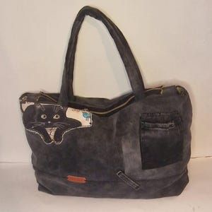 May include: A gray denim tote bag with a black cat graphic on the front. The bag has a zipper closure, a small pocket, and two handles. The cat graphic is on a patch with a white background.