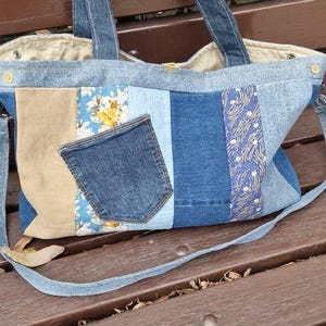 Peut inclure: Un sac fourre-tout en denim patchwork avec une variété de nuances de denim bleu, des accents beiges et floraux. Le sac comprend une poche en jean et une longue bandoulière réglable. Le sac est posé sur un banc en bois.