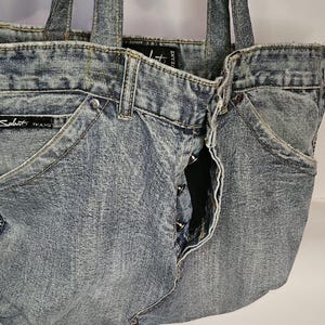 May include: A denim tote bag made from recycled jeans. The bag features the front pockets, button closure, and belt loops of the original jeans. The bag has two denim handles and the brand name "Sabit Jeans" is visible on the front.
