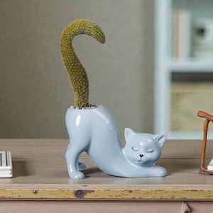 May include: A light blue ceramic cat-shaped planter with a cactus. The cat is in a stretching pose with its eyes closed. The cactus is green and curves upwards. The planter is on a wooden surface.