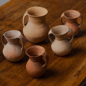 May include: Five small, handmade terracotta jugs with handles, arranged on a rustic wooden surface. The jugs vary in color from light beige to reddish-brown, showcasing a natural, earthy aesthetic. The vessels have a simple, classic design.